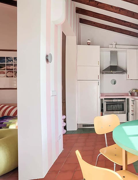 A bright kitchen area with a teal round dining table, exposed wooden ceiling beams, white cabinetry, and large windows overlooking the city.