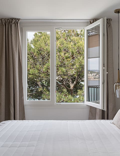 A bright bedroom with white linens, neutral curtains, and an open window overlooking pine trees and a distant coastline.