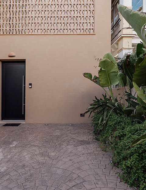 Entrance area of a modern townhouse featuring a dark door, beige stucco facade with patterned breeze block, and gravel ground.