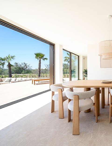 Dining room with light oak table and textured cream chairs, floor-to-ceiling glass walls opening to a sunlit terrace.