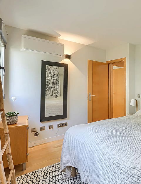 A bedroom featuring a double bed with a neutral headboard, textured white bedding, wooden door and cabinet, and wall-mounted air conditioning.