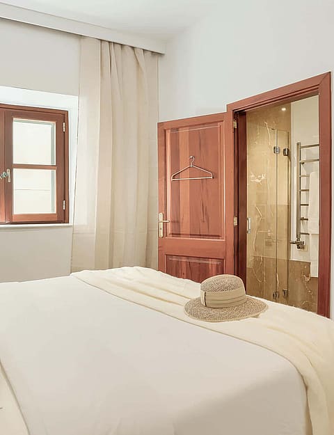 A bedroom interior with a white bed, beige curtains, a wooden window frame, and an open wooden door leading to a bathroom.