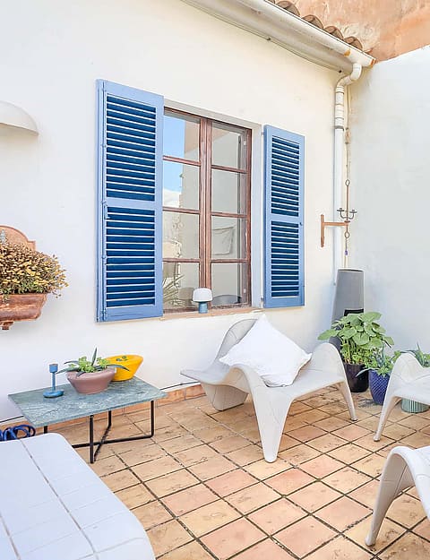 A small outdoor terrace with terracotta tiles, white plastered walls, blue wooden shutters, and modern white lounge chairs.