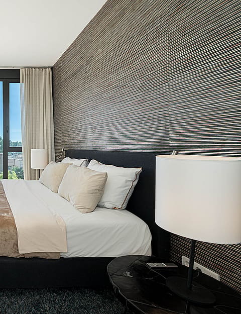 Modern master bedroom with king-sized bed, textured dark feature wall, and full-height windows overlooking a coastal scene.