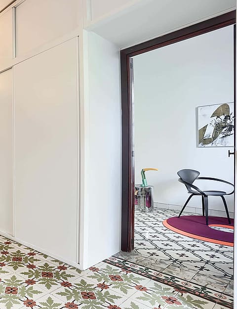 A bright hallway connecting two rooms, featuring patterned colorful tile flooring, white walls, and built-in storage closets.