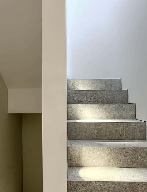 Interior staircase made of speckled grey stone with plain white walls and bright natural light from a skylight above.
