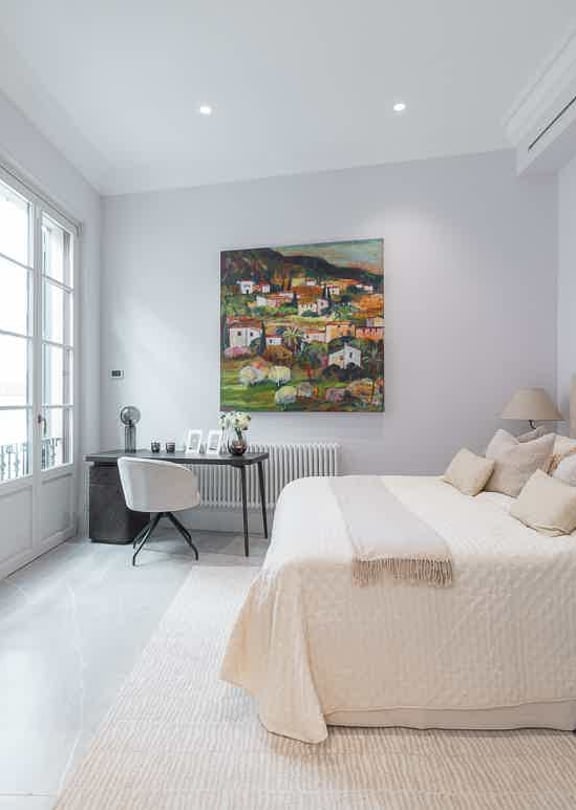 A minimalist bedroom featuring a double bed with neutral linens, a workspace by a window, and full-height mirrored wardrobe doors.