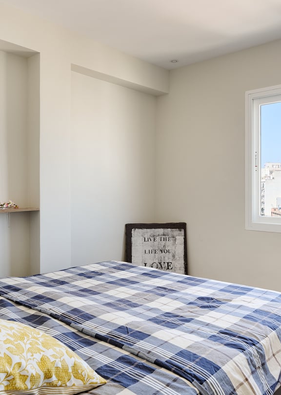 A bedroom featuring a bed with a blue plaid quilt, a floating wooden shelf with framed artwork, and a window overlooking city buildings.
