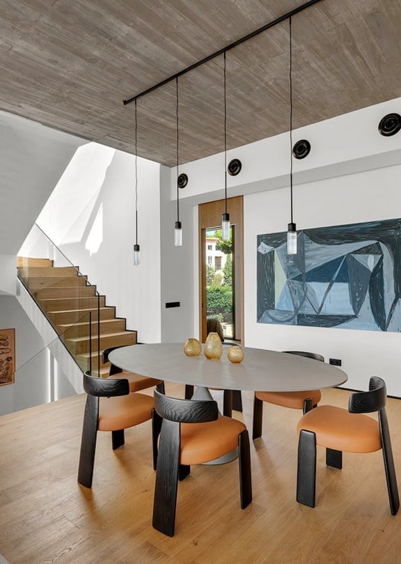 Dining area with oval table and wooden chairs, adjacent to a wooden staircase under a raw concrete ceiling.