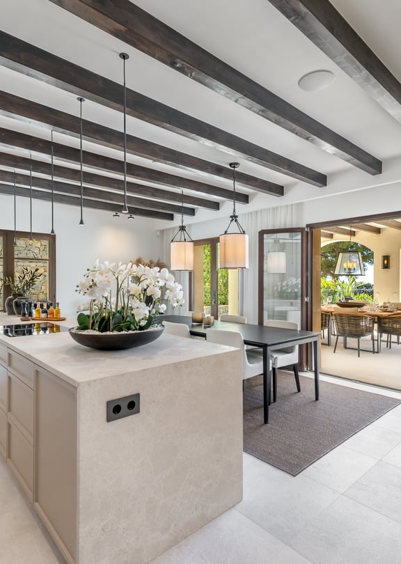 A wide stone kitchen island and dark ceiling beams in an open-plan room opening onto a sunlit outdoor dining terrace.