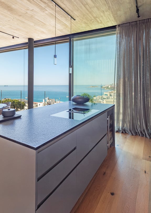 Modern kitchen island with dark stone countertop, light wood flooring, and floor-to-ceiling glass windows overlooking the sea.