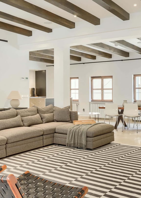 A spacious living room featuring a large patterned sofa, exposed dark wood ceiling beams, and a dining area in the background.