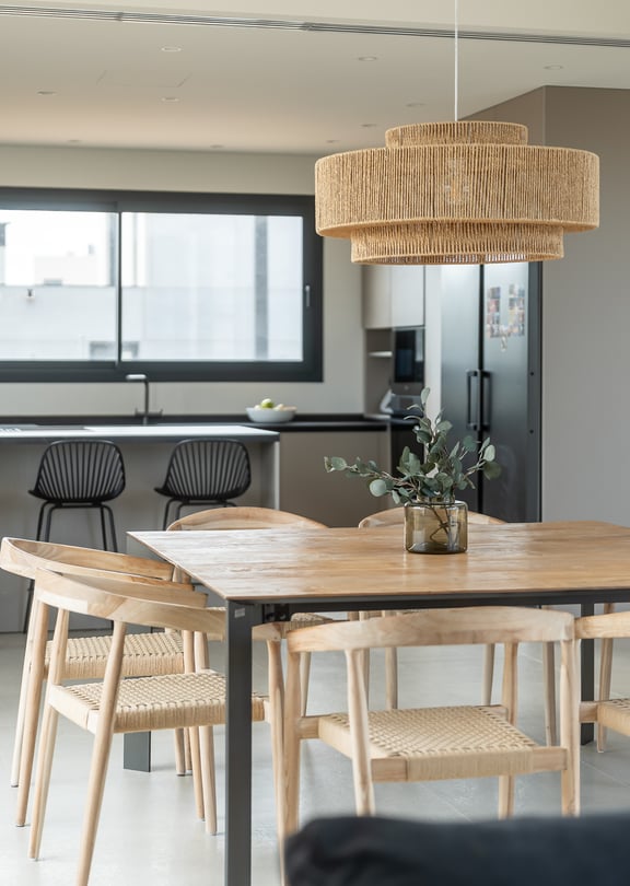 A dining space featuring a large square wooden table with woven chairs, a textured light fixture, and a modern kitchen island with black bar stools in the background.