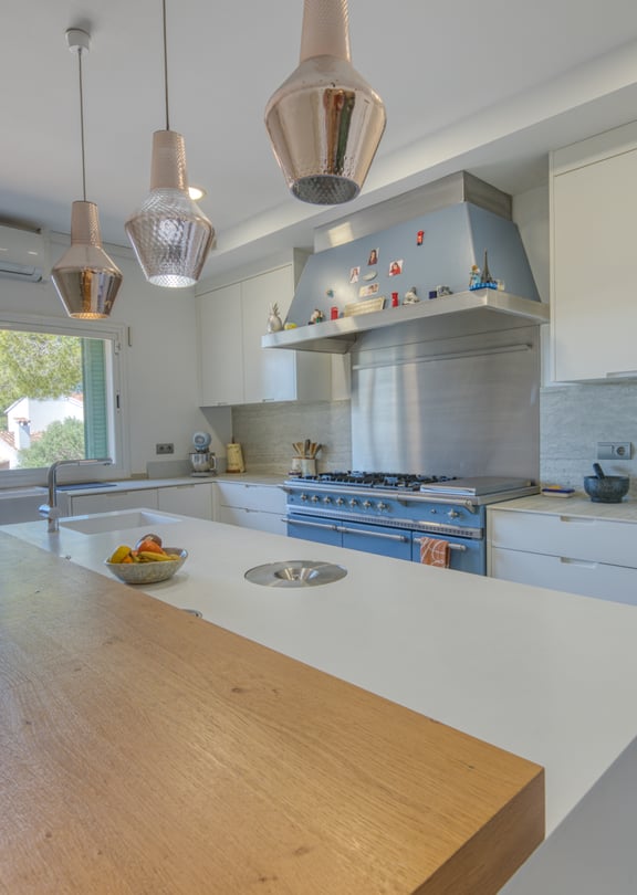 A modern white kitchen featuring a large central island with a wooden breakfast bar, stainless steel stove, and three glass pendant lights hanging above.