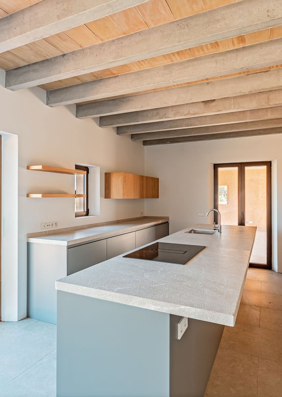 Modern kitchen featuring a central island, light grey cabinetry, wooden ceiling beams, and large glass doors overlooking a field.