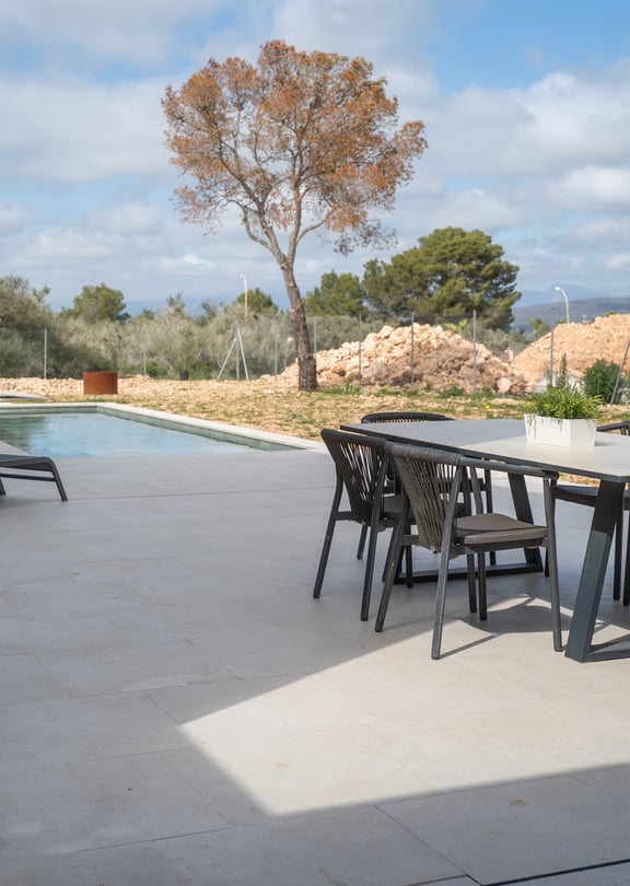 A covered terrace with a dining set and sun loungers overlooks a swimming pool and distant hills under a sunny sky.