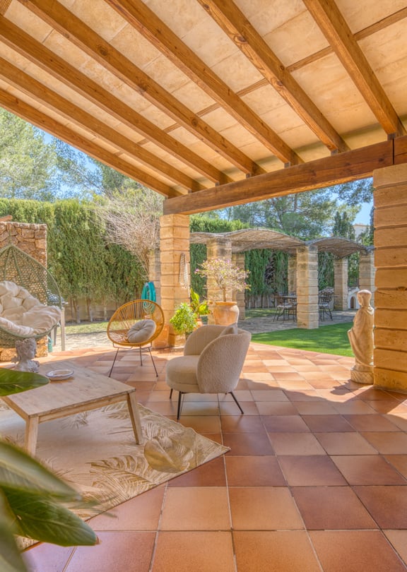 A covered terrace with terracotta floor tiles and wooden beam ceiling, featuring outdoor armchairs and a dry stone wall.