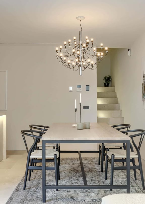 Interior view of a minimalist dining room with a stone table, black wooden chairs, a modern branching chandelier, and an abstract multi-colored painting on a white wall.