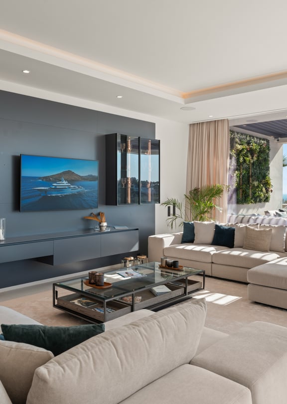 Modern living room with a charcoal grey feature wall, a wall-mounted flat-screen television, a beige sectional sofa, and floor-to-ceiling glass sliding doors opening to a terrace with sea views.