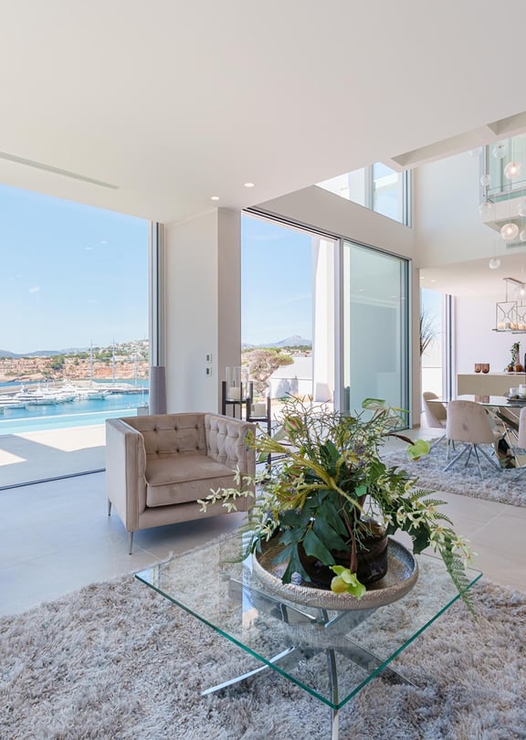 Modern open-plan living room with floor-to-ceiling windows overlooking Port Adriano marina, featuring tufted furniture.