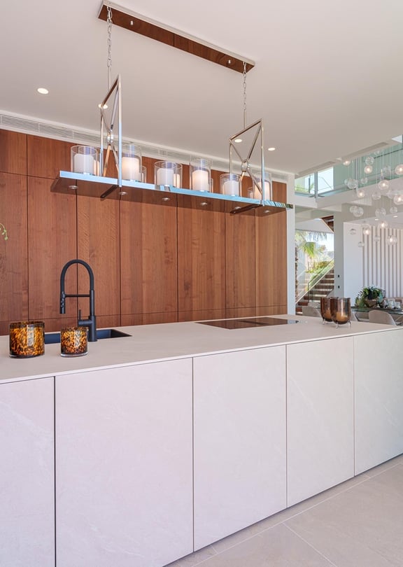 Modern kitchen island with matte white surfaces and dark wood wall-to-ceiling cabinetry, interior glass staircase visible in background.