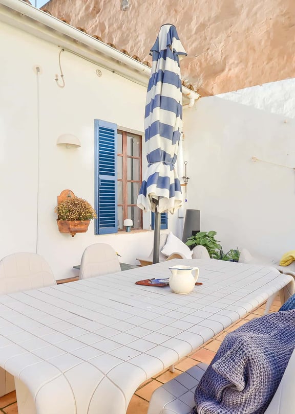 Outdoor terrace with a dining table and chairs, blue-striped sun umbrella, and blue shutters on a white building exterior.