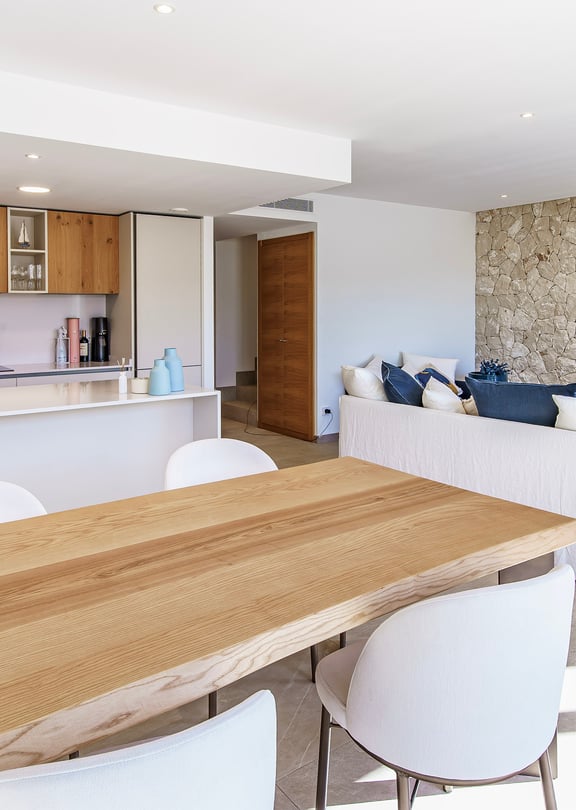 A bright open-plan space showing a kitchen with wooden cabinetry, a breakfast bar, and a living area featuring a stone wall.