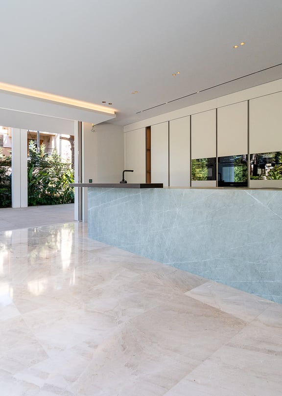 Modern kitchen featuring a large grey stone island, handleless white cabinets, and polished light marble floors. A wide glass door opens to a stone terrace and garden.