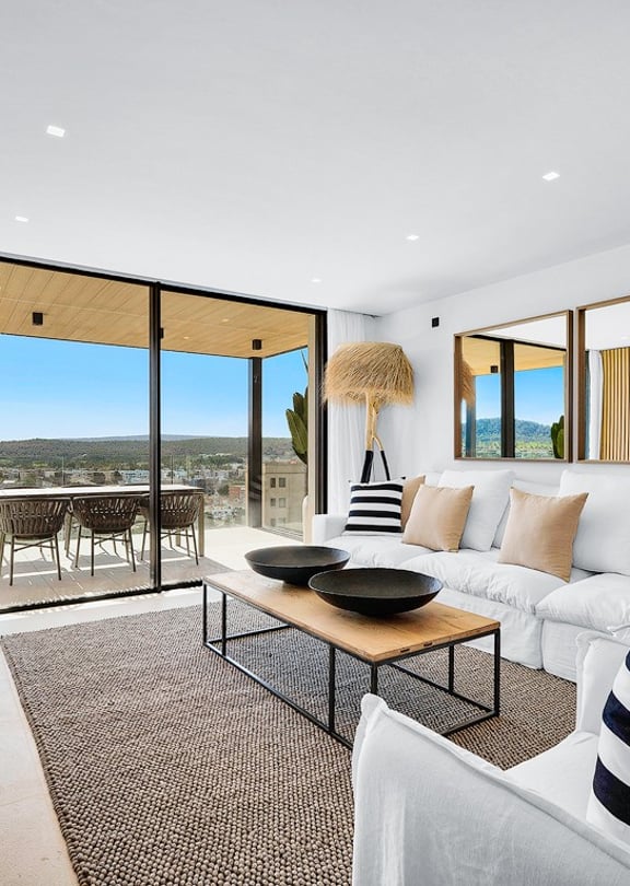 Modern open-plan living room with white sofas, wooden wall paneling, and large glass doors opening onto a terrace with mountain views.