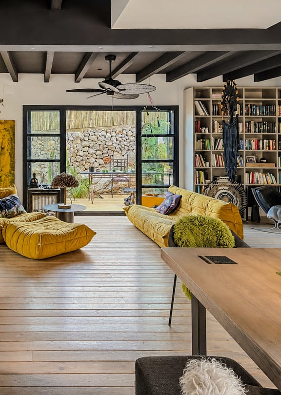 A living area with light wood floors, a large yellow sofa, and a wall-to-wall integrated bookshelf opening onto a stone terrace.
