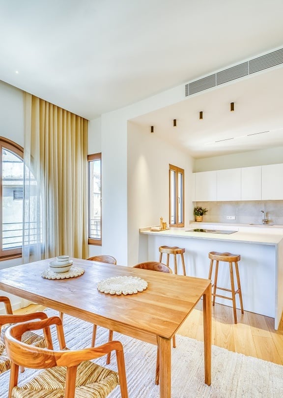 A bright open-plan kitchen and dining room with wooden furniture, white cabinetry, and large arched windows draped in curtains.