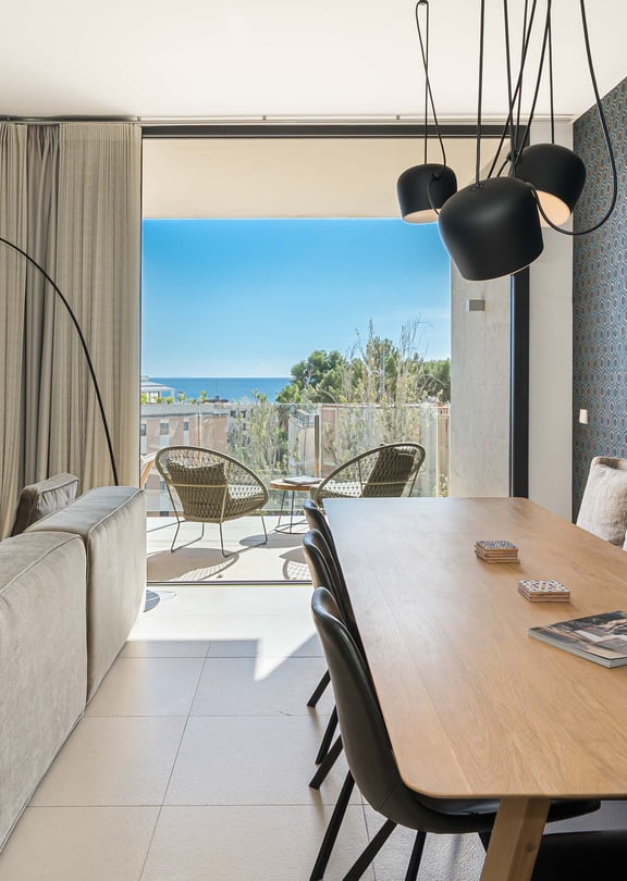 Interior view of an open-plan living and dining room with floor-to-ceiling glass doors opening onto a terrace with a sea view.