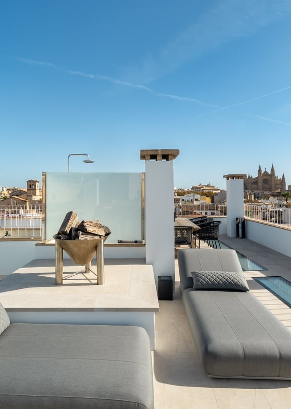 Sun-drenched rooftop terrace with grey loungers, stone flooring, and panoramic views of Palma's historic skyline.