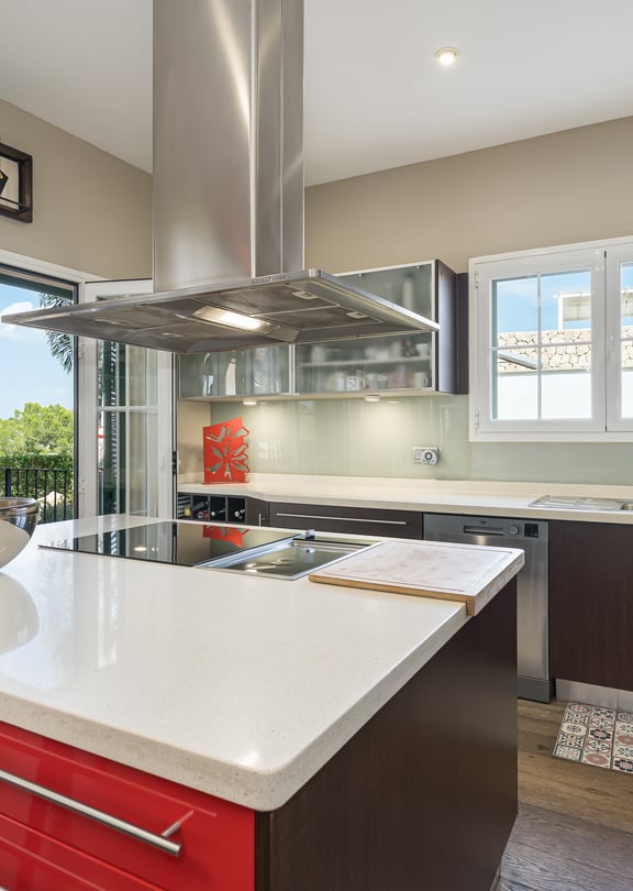 A modern kitchen featuring a bold red central island with a white stone countertop, dark cabinetry, and floor-to-ceiling glass doors opening to a terrace.