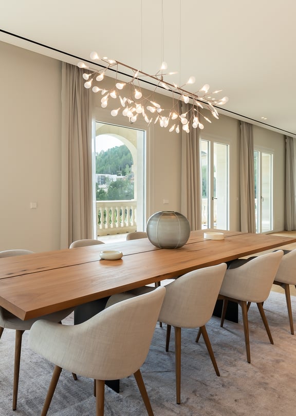Dining room featuring a long wooden table, light-colored upholstered chairs, and a sculptural chandelier, overlooking the sea.