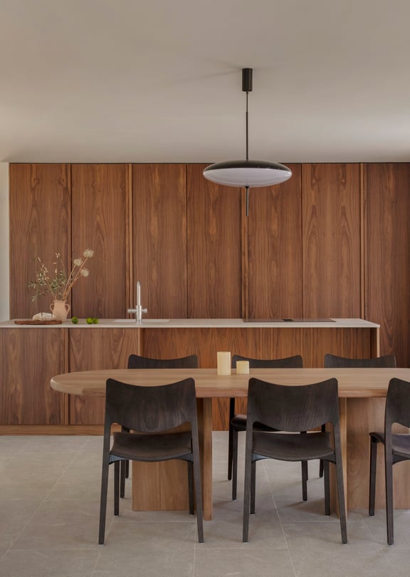 A minimalist dining area featuring an oval wood table and dark chairs, set against full-height walnut kitchen cabinetry.