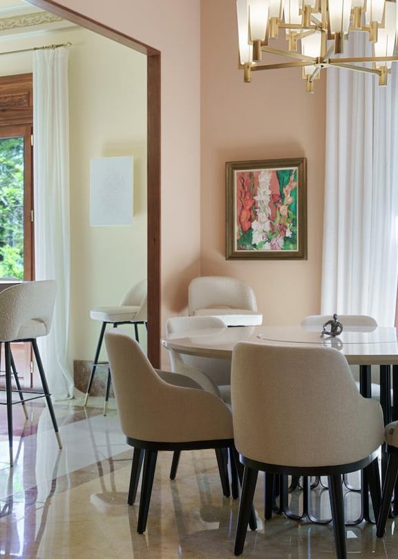 Dining room with a round marble table, upholstered chairs, a crystal chandelier, and large windows overlooking trees.