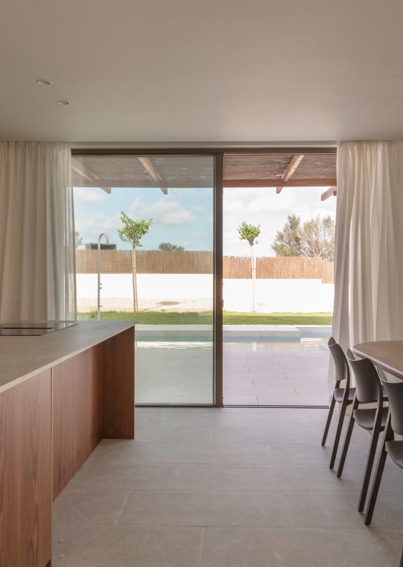 An oval oak dining table and dark chairs in a sunlit room with stone floors and floor-to-ceiling glass sliding doors.