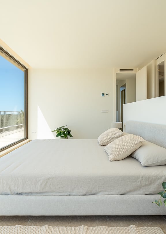 Modern bedroom with a double bed, floor-to-ceiling glass wall, white walls, and view of the exterior landscape.