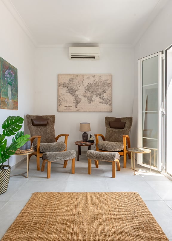 A bright living space with two armchairs, footstools, a woven rug, and floor-to-ceiling glass doors opening to a terrace.