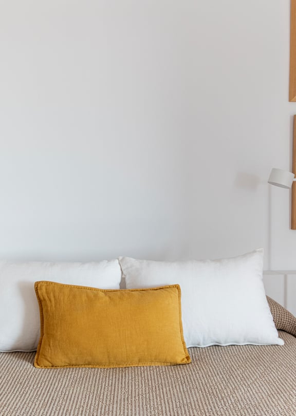 A double bed with a patterned beige cover and mustard yellow cushion, flanked by two minimalist floor lamps and framed wall art.