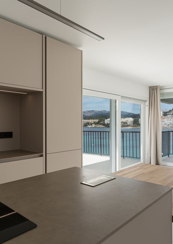Modern taupe kitchen and wooden island extension facing floor-to-ceiling windows with a view of a coastal harbor.