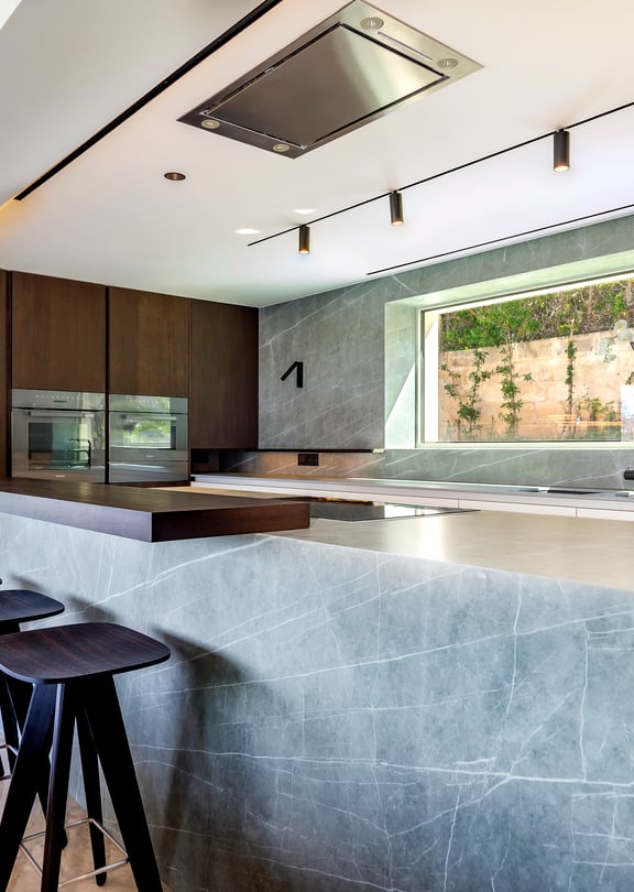 Modern kitchen with a large grey marble island, wooden cabinets, and three dark wooden bar stools under track lighting.