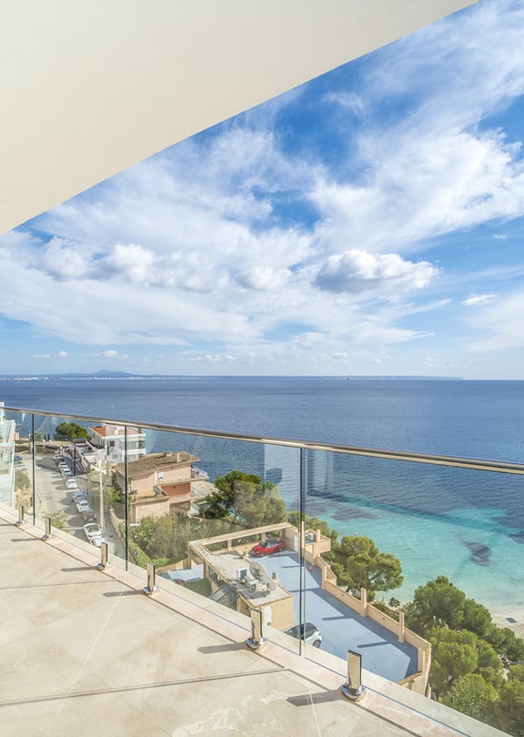 A wide sun-drenched terrace overlooking a turquoise Mediterranean bay with a glass railing and outdoor seating.