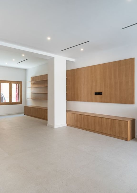An expansive, bright living space with large light-grey floor tiles, custom light oak cabinetry, and a black metal fireplace.