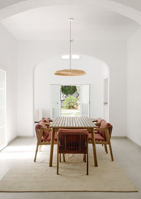 Comedor con bóveda blanca, mesa de teca, sillas de cuerda y lámpara de ratán, vistas al jardín a través de puertas abiertas