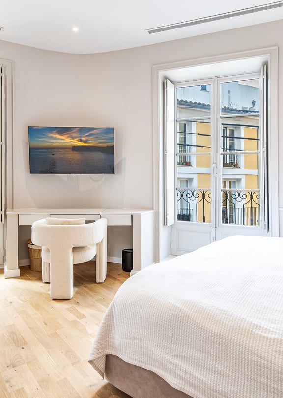 Bright bedroom with white walls, light wood floors, a desk, and floor-to-ceiling windows looking out onto a yellow building.