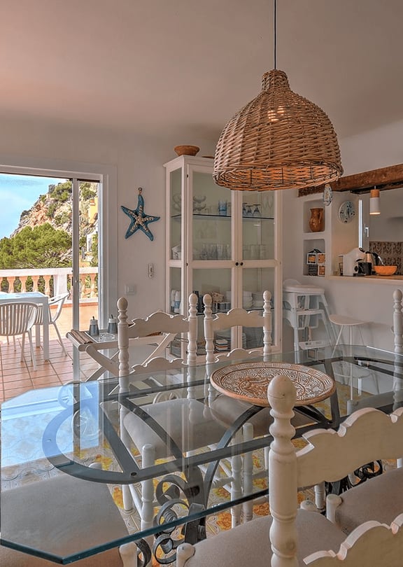 Interior dining room with a glass-top table, white wooden chairs, and a rattan pendant light, opening to a terrace with sea views.