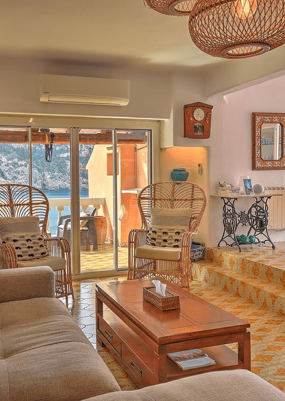 Sunlit living area with tiled floors, rattan armchairs, and a view of Cala Llamp through glass sliding doors.