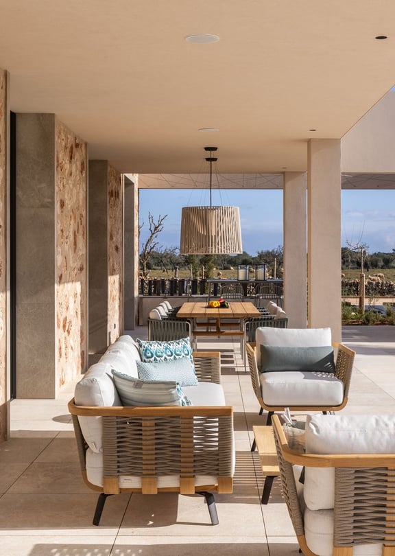 A spacious covered terrace with stone walls, outdoor lounge seating, and a dining area, overlooking a distant mountain landscape.